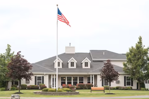 Front view of a single-story residential building with an American flag on a pole, landscaped lawn, and a bench.
