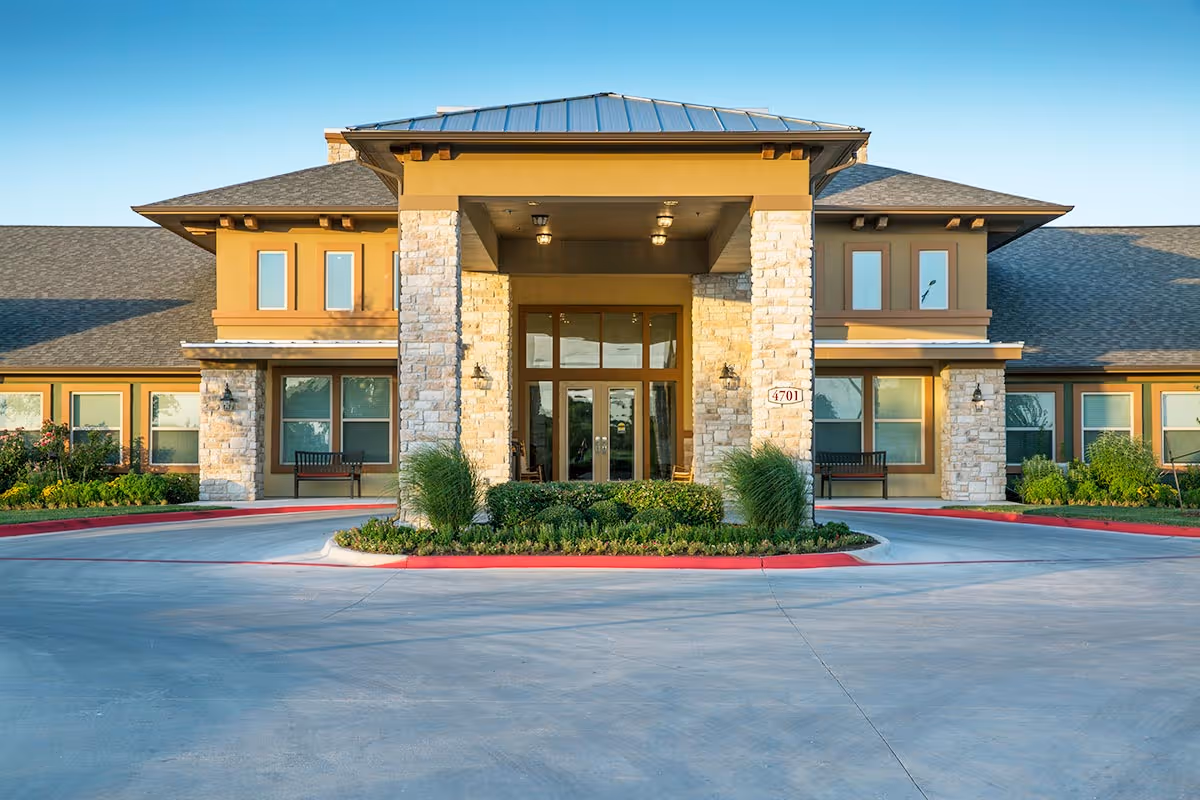 Front exterior view of Orchard Park of Kyle Assisted & Senior Living facility showing a large entrance with stone pillars, glass doors, and surrounding windows. There are benches on either side of the entrance and landscaped greenery in front.