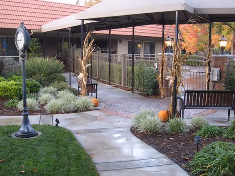 Covered courtyard walkway with benches, landscaping, pumpkins, and a decorative clock outside a senior living building.