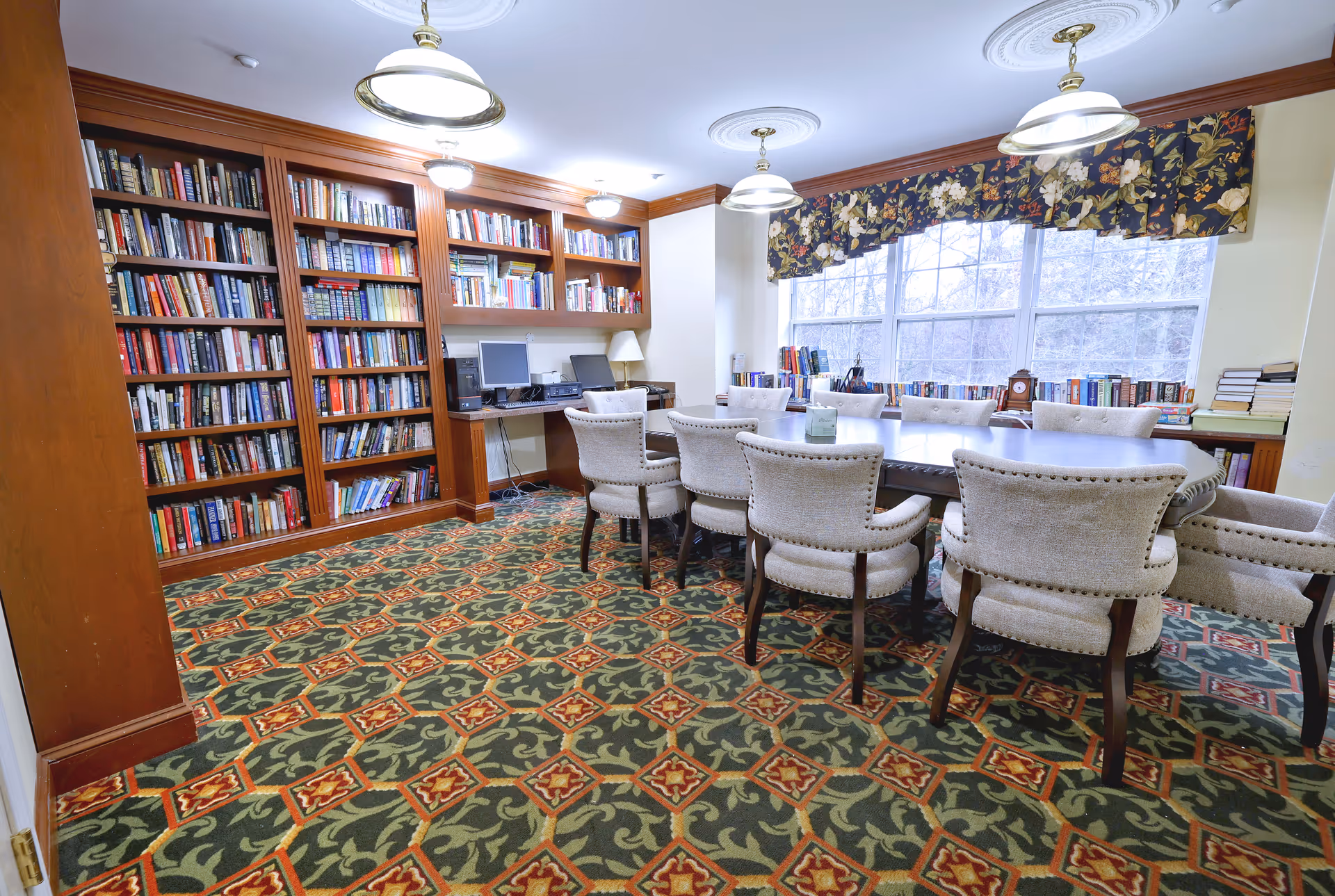 A well-lit room with a large wooden table surrounded by upholstered chairs. The room features a patterned carpet, built-in wooden bookshelves filled with books, and a desk area with two computers. Large windows with floral valances allow natural light to enter the space.