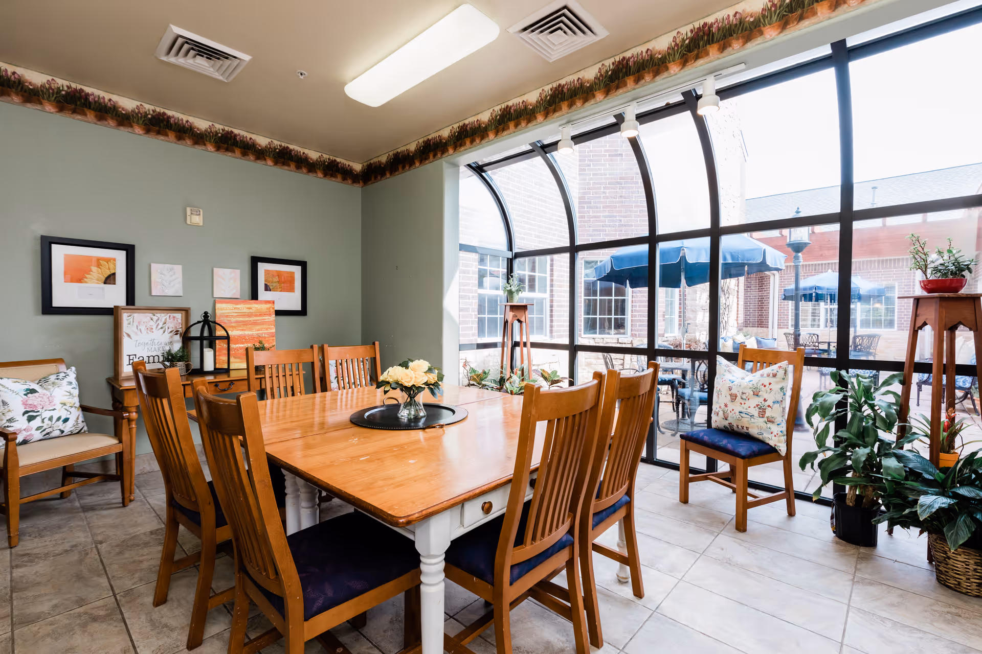 A bright dining area with a wooden table surrounded by eight wooden chairs with blue cushions. The room has large curved windows letting in natural light and showing an outdoor patio with tables and blue umbrellas. The walls are painted light green with floral border trim near the ceiling. There are framed pictures and decorative items on the walls and furniture, along with several potted plants around the room.