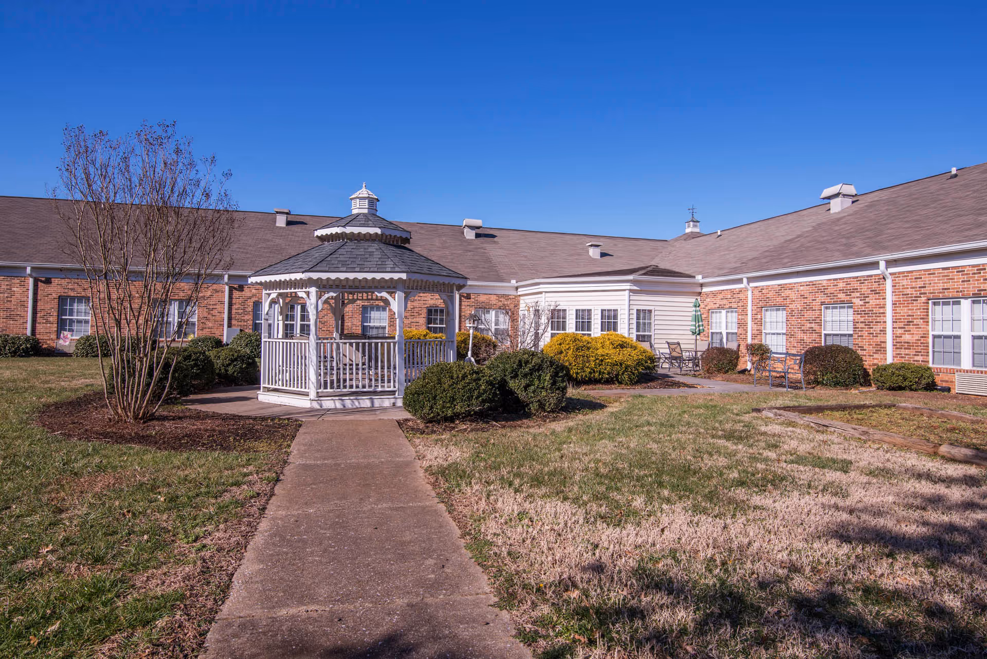 A brick senior living facility courtyard with a white gazebo centered on a paved path, surrounded by lawn, shrubs and a clear blue sky.