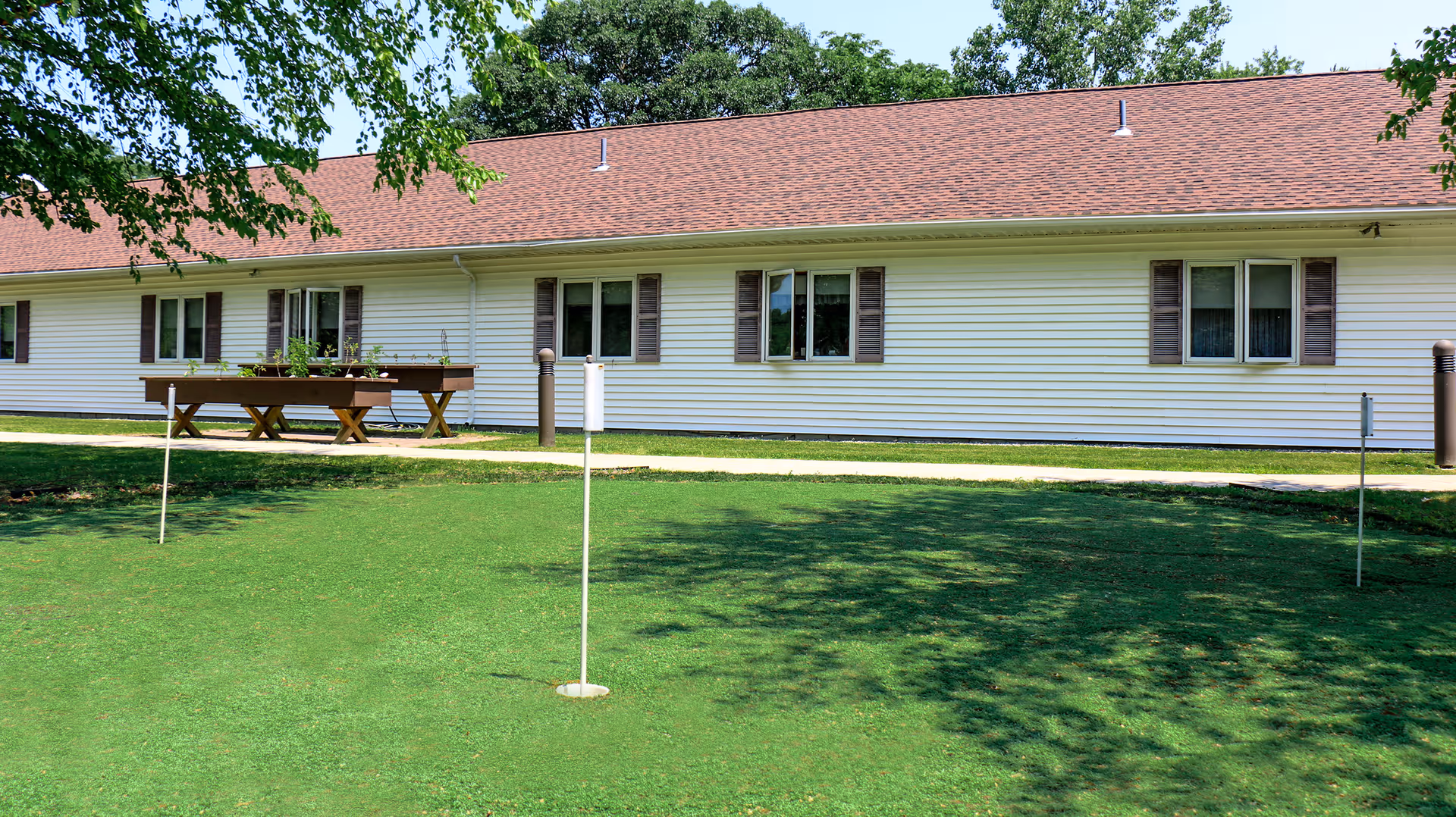 Outdoor view of a single-story building with white siding and brown shutters, surrounded by green grass and trees. There are several small flag poles on the grass, possibly indicating a putting green area.
