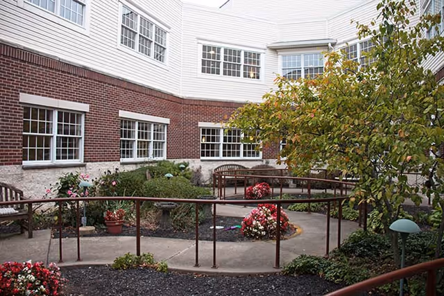 Small landscaped courtyard with benches, flower beds, a wheelchair ramp, and windows of a surrounding brick-and-siding building.