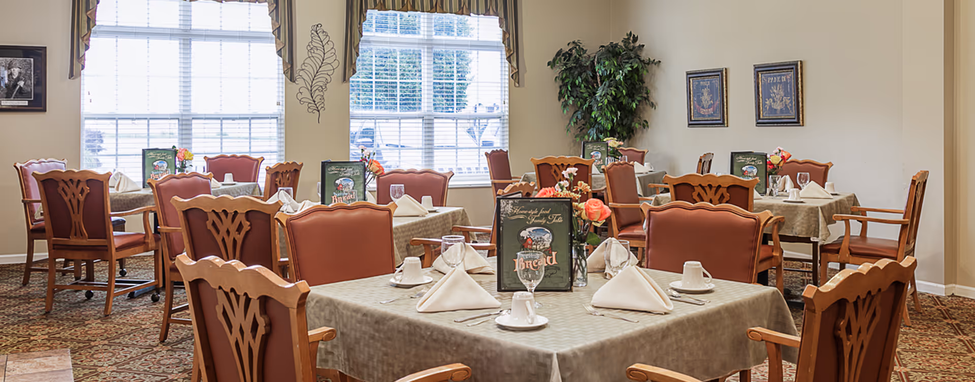 Bright dining room with multiple tables set with napkins, plates, glassware and floral centerpieces, surrounded by wooden chairs and large windows.
