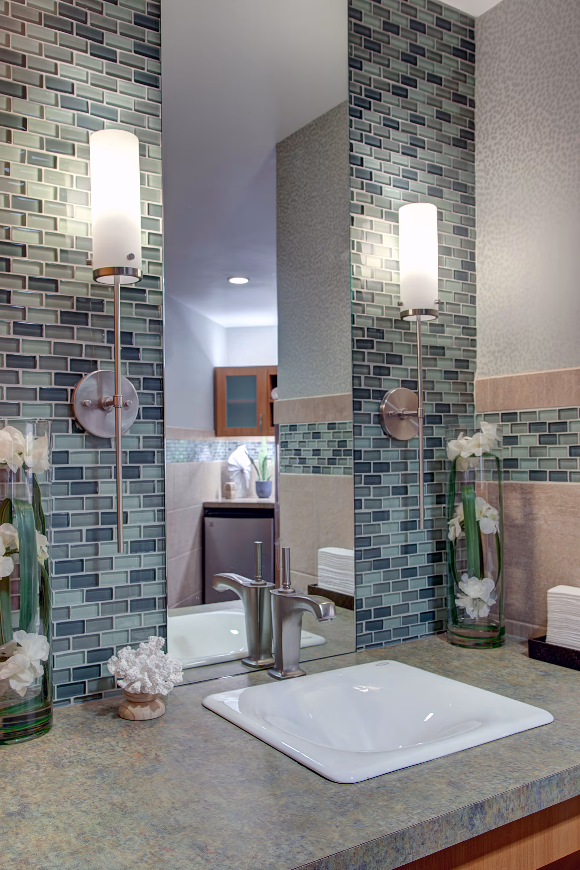 A bathroom sink area with a modern faucet, a large mirror, and two wall-mounted cylindrical light fixtures. The wall behind the sink is decorated with green and blue mosaic tiles. There are two glass vases with white flowers on either side of the sink, and a stack of white napkins or towels on the right side of the counter.
