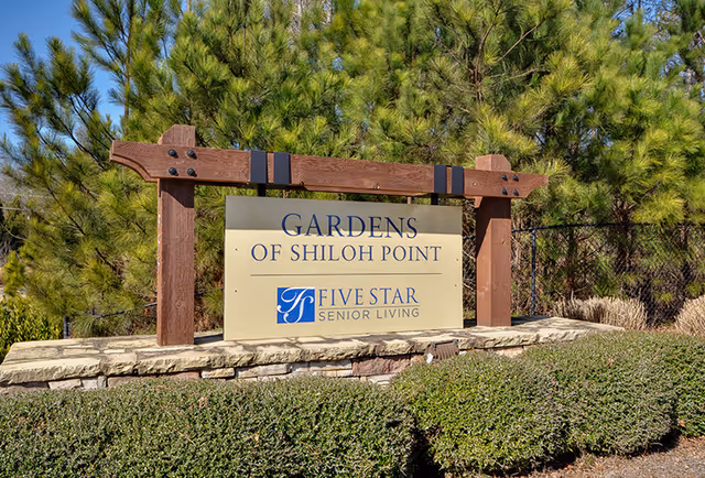 Outdoor sign for Gardens of Shiloh Point, a Five Star Senior Living community, mounted on a stone base with wooden posts, surrounded by green shrubs and pine trees in the background under a clear blue sky.