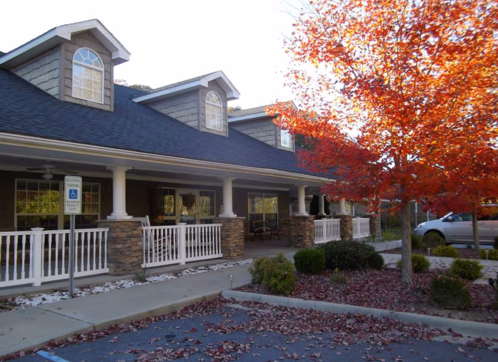 Exterior view of Hayesville House showing a building with a covered porch supported by white columns and stone bases. There are large windows and a dark shingled roof with dormer windows. In front, there is a parking area with a reserved handicapped parking sign, a tree with bright red autumn leaves, and some landscaping with bushes and fallen leaves on the ground.