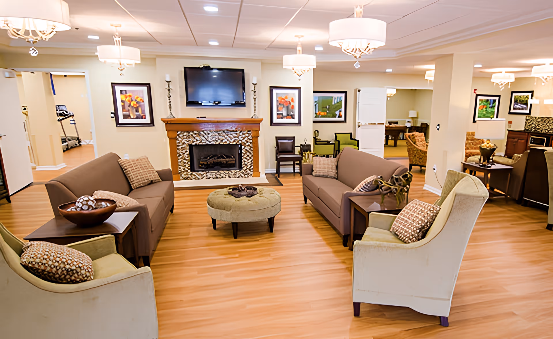 A cozy senior living facility common area with two brown sofas, two beige armchairs with patterned cushions, a round green ottoman in the center, and a fireplace with a mounted flat-screen TV above it. The room has wooden flooring, soft lighting from ceiling fixtures, and framed artwork on the walls. In the background, there are additional seating areas and a glimpse of exercise equipment in an adjacent room.