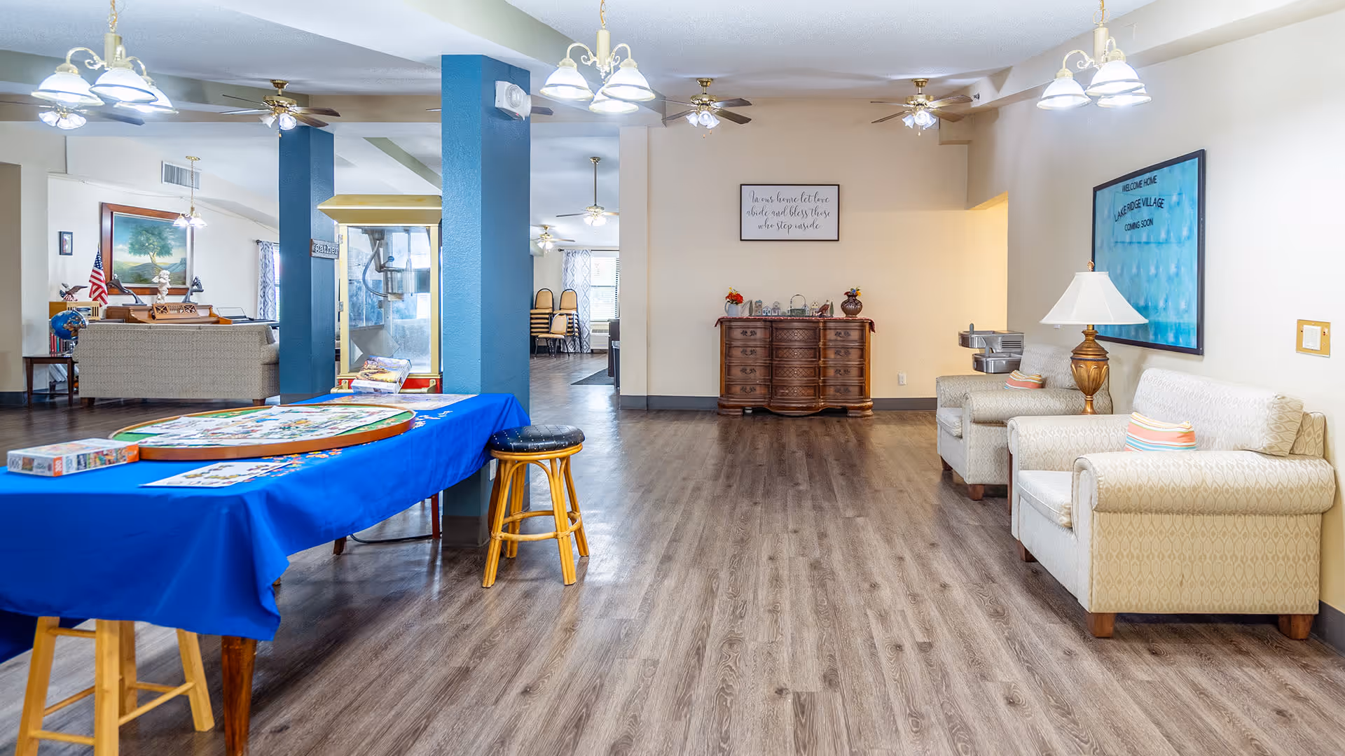 Interior view of a senior living facility common area with wood flooring, beige walls, and ceiling fans. There are two beige armchairs with a lamp and a water fountain on the right side. A wooden dresser with decorative items is against the far wall. A blue tablecloth covers a table with a puzzle and puzzle pieces on it. In the background, there is a seating area with a couch, an American flag, and a large painting of a tree. A sign on the wall reads 'Welcome Home Lake Ridge Village Coming Soon.'