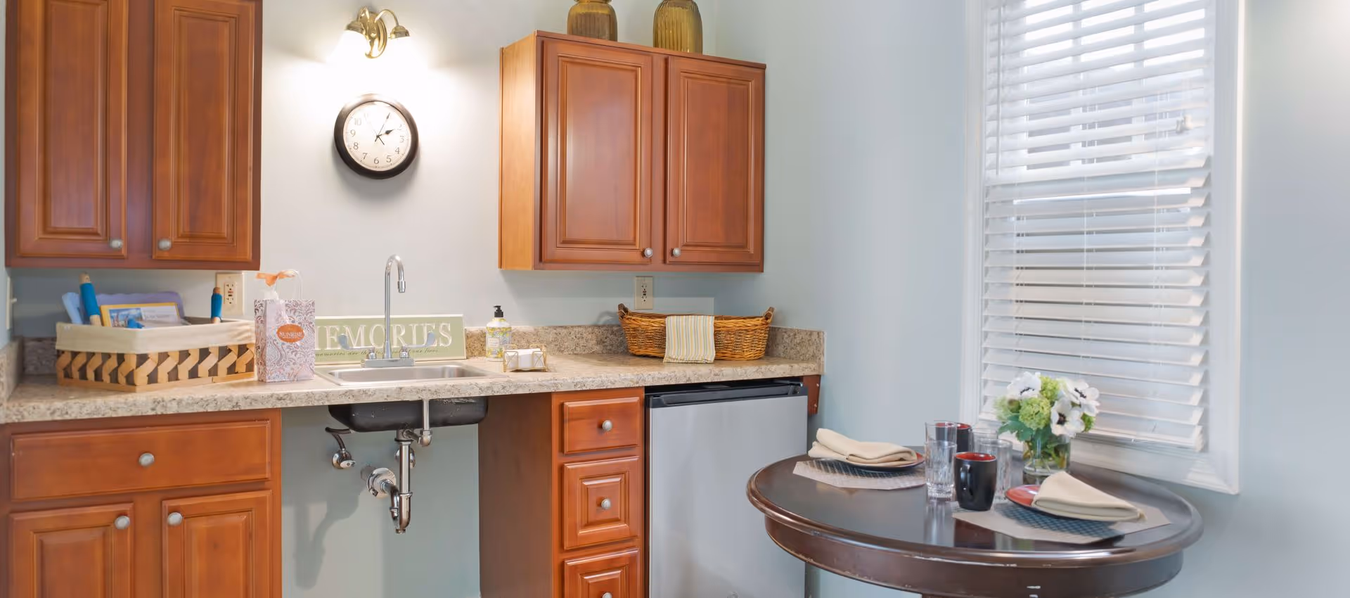 A small kitchen area with wooden cabinets, a sink, a wall clock, and a countertop with a basket and soap dispenser. Next to the kitchen is a round dining table set with two place settings, glasses, mugs, and a small vase with flowers. A window with white blinds is behind the table.