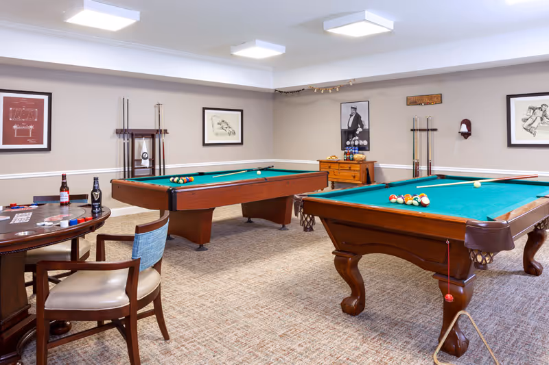 A recreational room with two wooden pool tables with green felt tops, pool cues resting on racks on the walls, and a small wooden cabinet with billiard balls and accessories. There is also a round table with chairs, poker chips, and two bottles of beer. The walls are decorated with framed pictures and the room is well-lit with ceiling lights.
