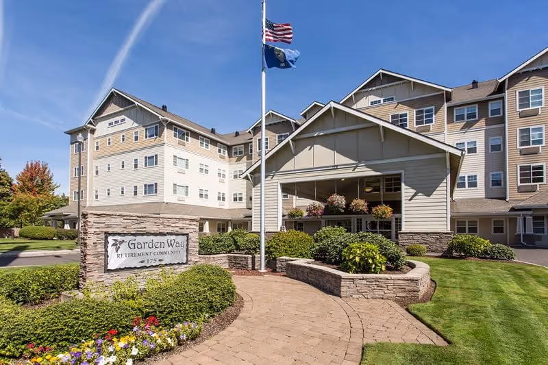 Front entrance of the Garden Way Retirement Community building with flagpoles, landscaped walkways, and a stone sign.