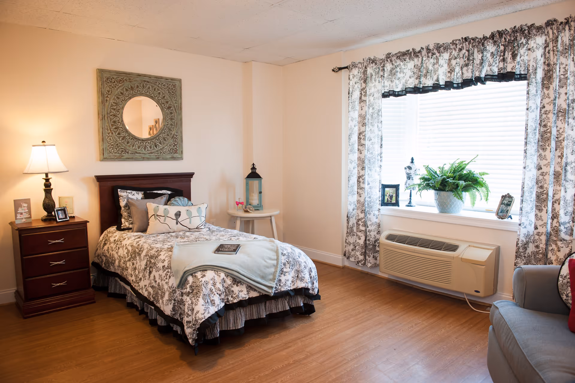 A cozy bedroom with a single bed covered in a floral patterned bedspread and decorative pillows. There is a wooden nightstand with a lamp and picture frames on the left side of the bed. A round side table with a lantern and small decorative items is next to the bed. A large window with floral curtains lets in natural light, and a potted plant and picture frames are on the windowsill. A gray armchair is partially visible on the right side of the image. The floor is wooden, and a decorative round mirror hangs on the wall above the bed.