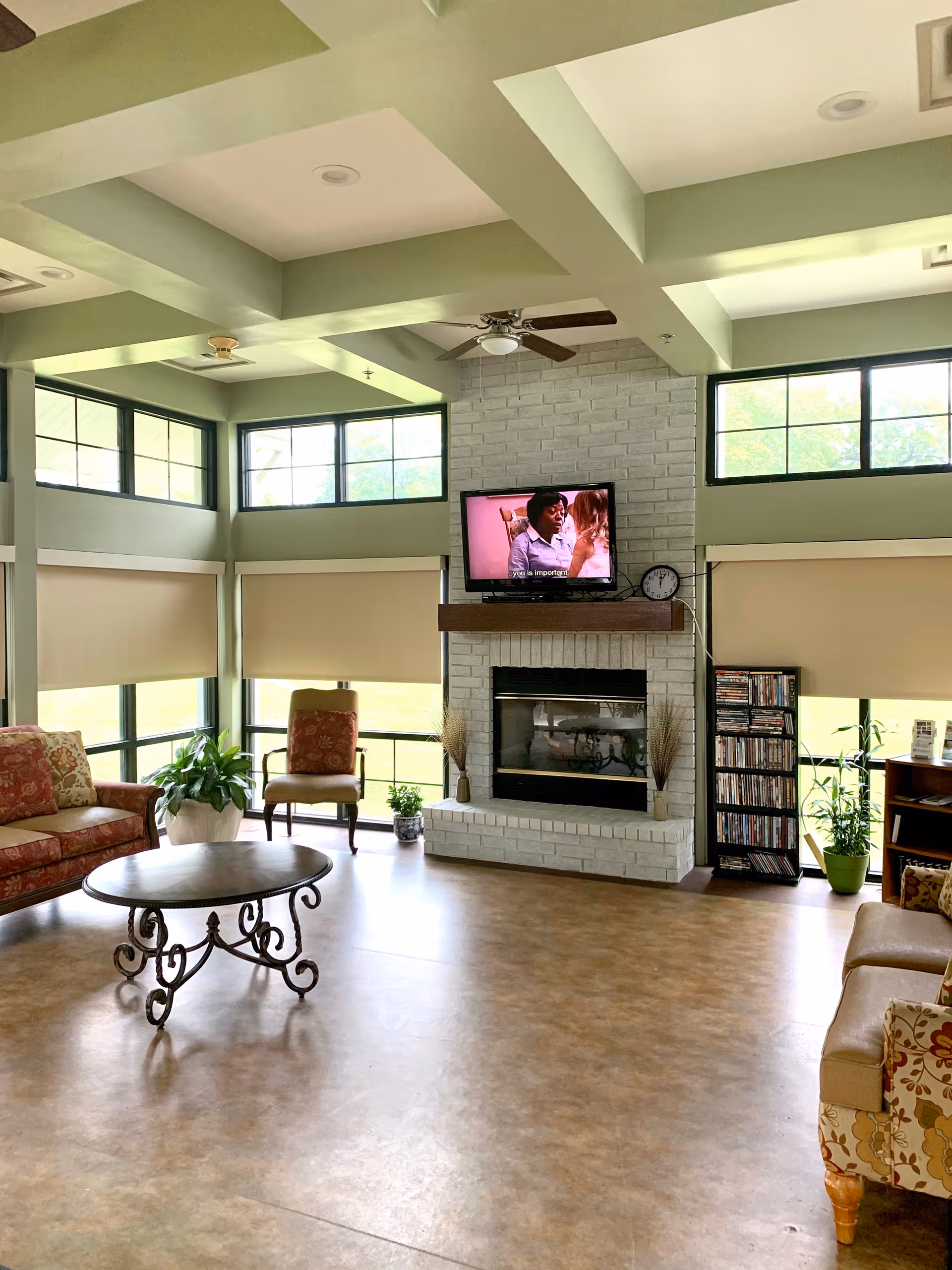 A bright communal living room with a coffered ceiling, large windows, a fireplace with a TV above, sofas, chairs, and a round coffee table.