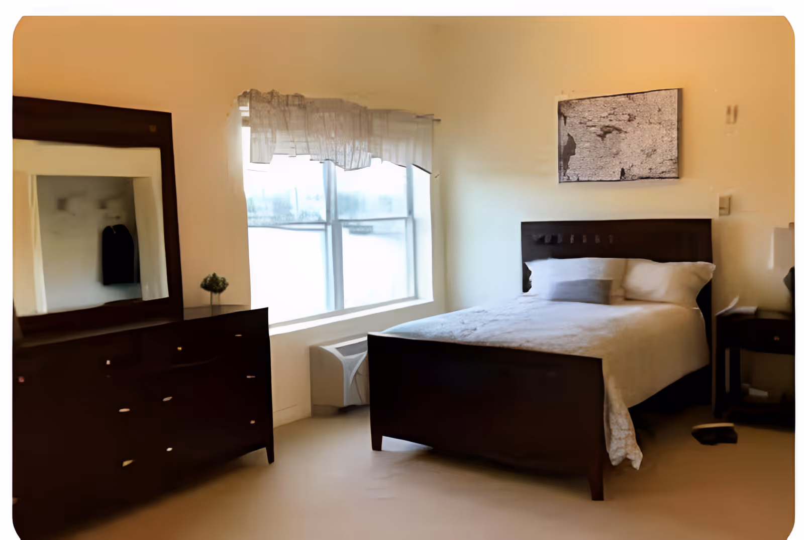 A bedroom with a dark wooden bed frame and a neatly made bed with white bedding and pillows. There is a matching dark wooden dresser with a large mirror on the left side of the room, and a small plant on top of the dresser. A window with sheer curtains allows natural light to enter the room. A piece of abstract artwork hangs on the wall above the bed. The room has a calm and simple decor.
