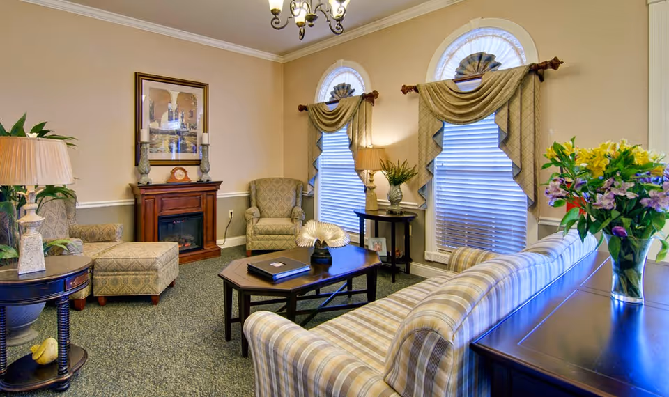 Cozy living room with plaid sofa, armchairs, coffee table, fireplace, and arched windows with draped valances.
