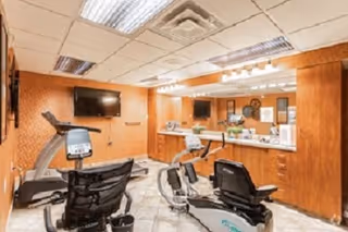 Indoor exercise room with two recumbent stationary bikes and a treadmill. The room has wood-paneled walls, a tiled floor, a large mirror, and two wall-mounted flat-screen TVs. The ceiling has fluorescent lighting panels.