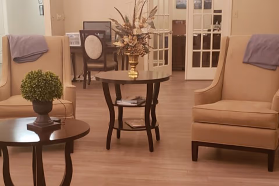 A cozy living room area with two beige armchairs, each draped with a purple throw blanket. Between the chairs are two dark wooden tables, one with a small green potted plant and the other with a decorative floral arrangement. In the background, there is a desk with a chair and French doors leading to another room.