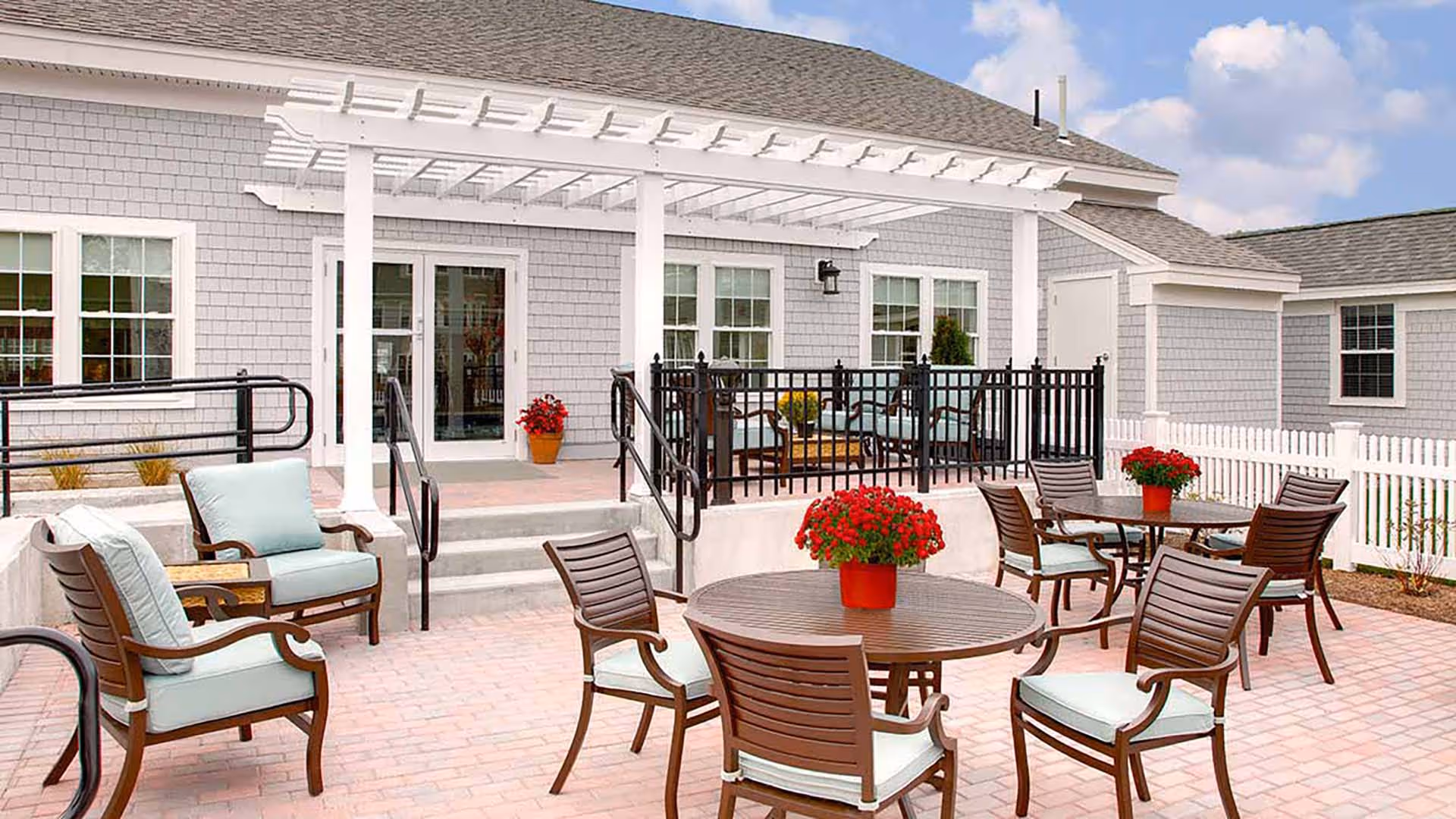 Outdoor patio area at Atria Woodbriar Park with multiple wooden tables and chairs featuring light blue cushions. There are red flower pots on the tables and a white pergola attached to the building. The building exterior is gray with white trim and multiple windows.