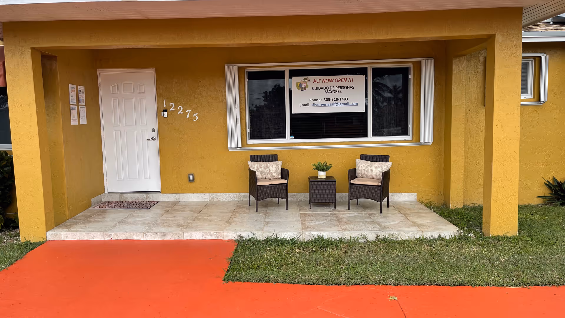 Front porch of a yellow building with two wicker chairs and a small table beneath a window displaying an 'ALF NOW OPEN' sign.