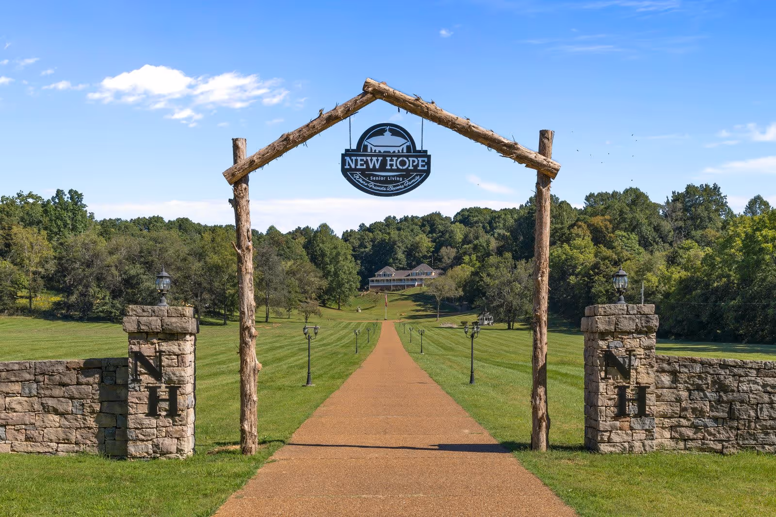 A long paved driveway passes under a rustic wooden arch with a hanging 'New Hope' sign leading to a large building set among lawns and trees.