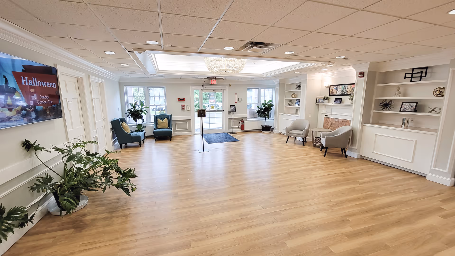 Bright, spacious lobby-style seating area with hardwood floors, chairs, built-in shelves, plants, and double glass doors.
