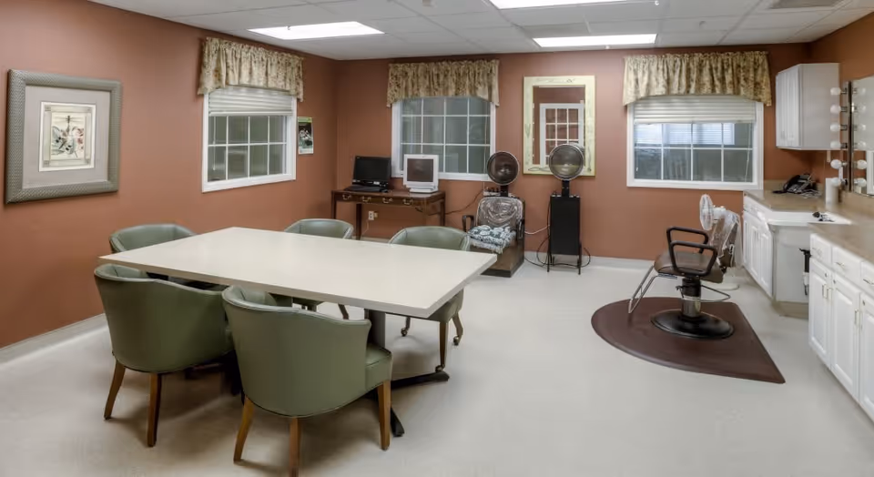 A room with a large rectangular table surrounded by six green chairs, two windows with floral valances, a hair salon chair on a brown mat in front of a counter with a sink and mirror with lights, two hair dryers on stands, and a small desk with two computers against a brown wall.