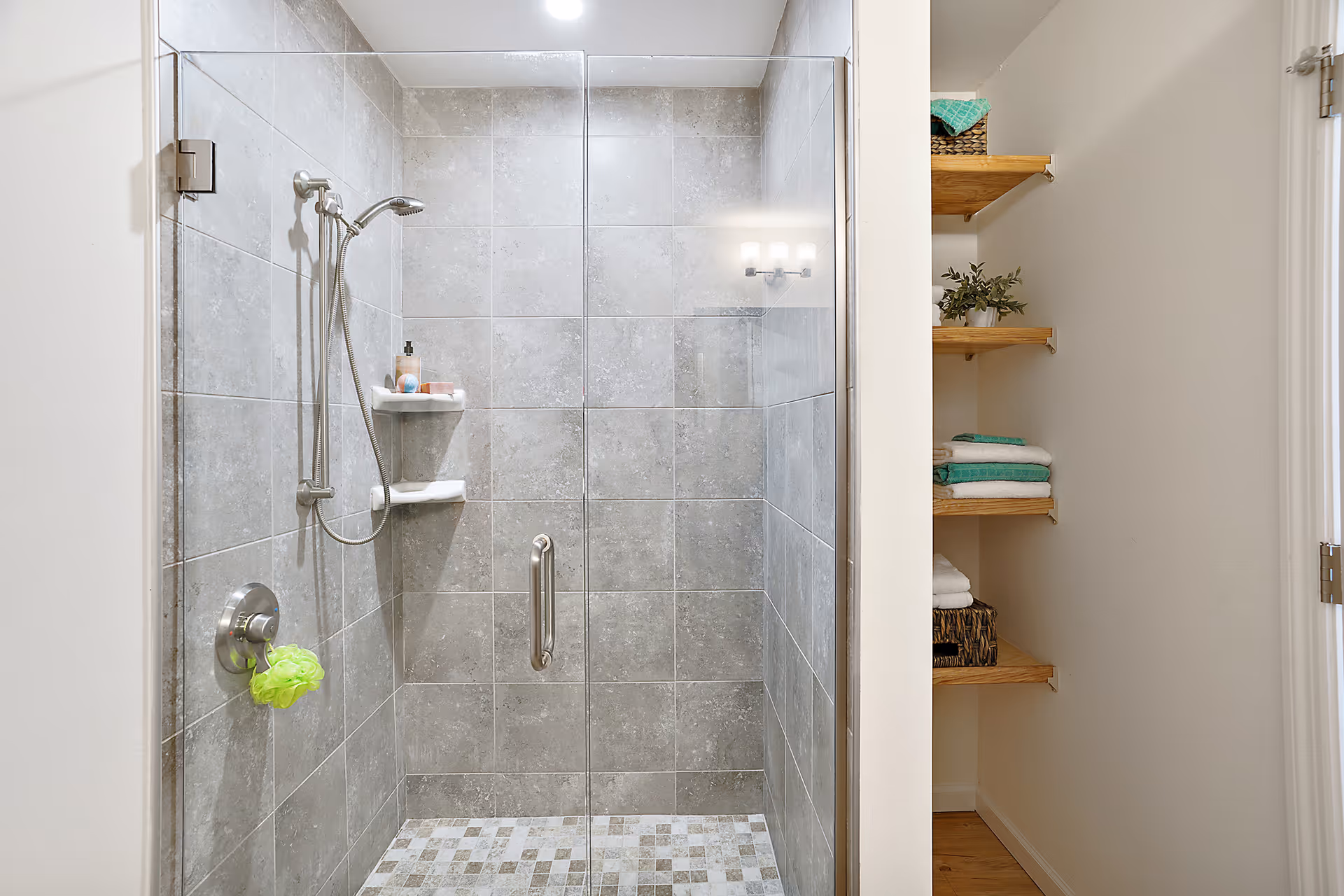A modern bathroom shower with gray tiled walls and floor, a glass door with a metal handle, a handheld showerhead, and built-in corner shelves holding soap and shampoo. To the right, wooden shelves hold folded towels and a small plant.