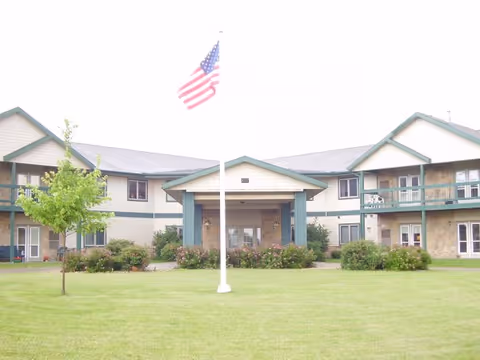 Exterior view of Stonefield Apartment Homes & Health Services building with a large green lawn in front, a flagpole with an American flag, and some bushes and small trees near the building.