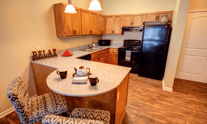 A cozy kitchen area with wooden cabinets, a black refrigerator, black stove, and a microwave. The kitchen has a light-colored countertop with a teapot and two mugs placed on it. Two patterned chairs are positioned at the counter. The word 'RELAX' is displayed on the countertop near the wall. The floor is wooden, and the walls are painted a light beige color.