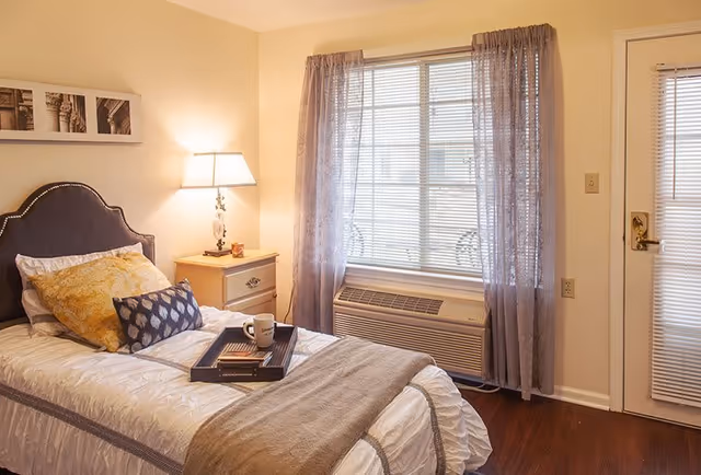 A cozy bedroom with a single bed made up with white and beige bedding, decorative pillows, and a tray holding a coffee mug and a book. Next to the bed is a nightstand with a lamp and a small candle. The room has a window with sheer curtains and an air conditioning unit below it, and a door with blinds on the right side.