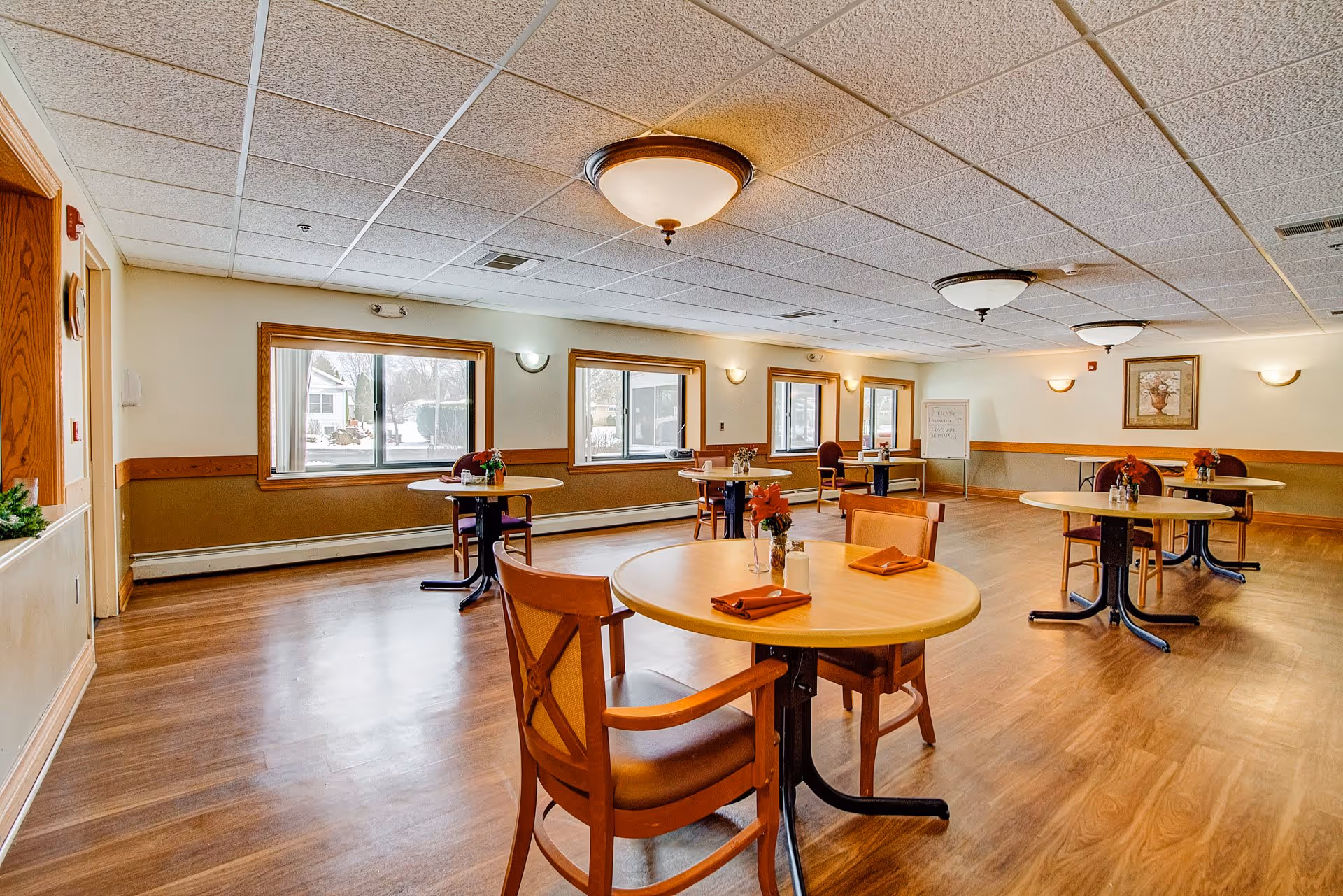 Well-lit communal dining room with round tables and chairs arranged on wood floors and place settings.