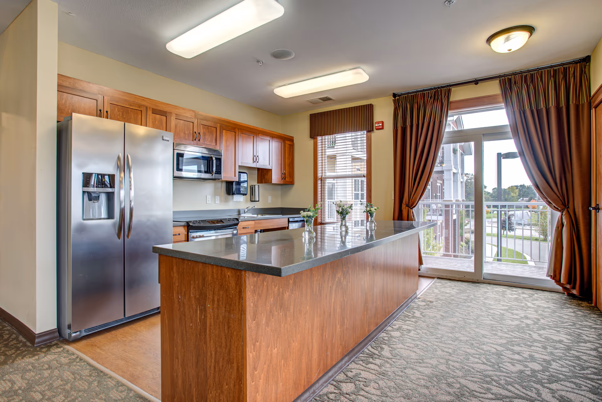 A modern kitchen area in a senior living facility featuring wooden cabinets, a stainless steel refrigerator, microwave, stove, and dishwasher. There is a large kitchen island with a dark countertop and three small vases with flowers on it. The room has large windows with brown curtains and a glass door leading to an outdoor balcony.