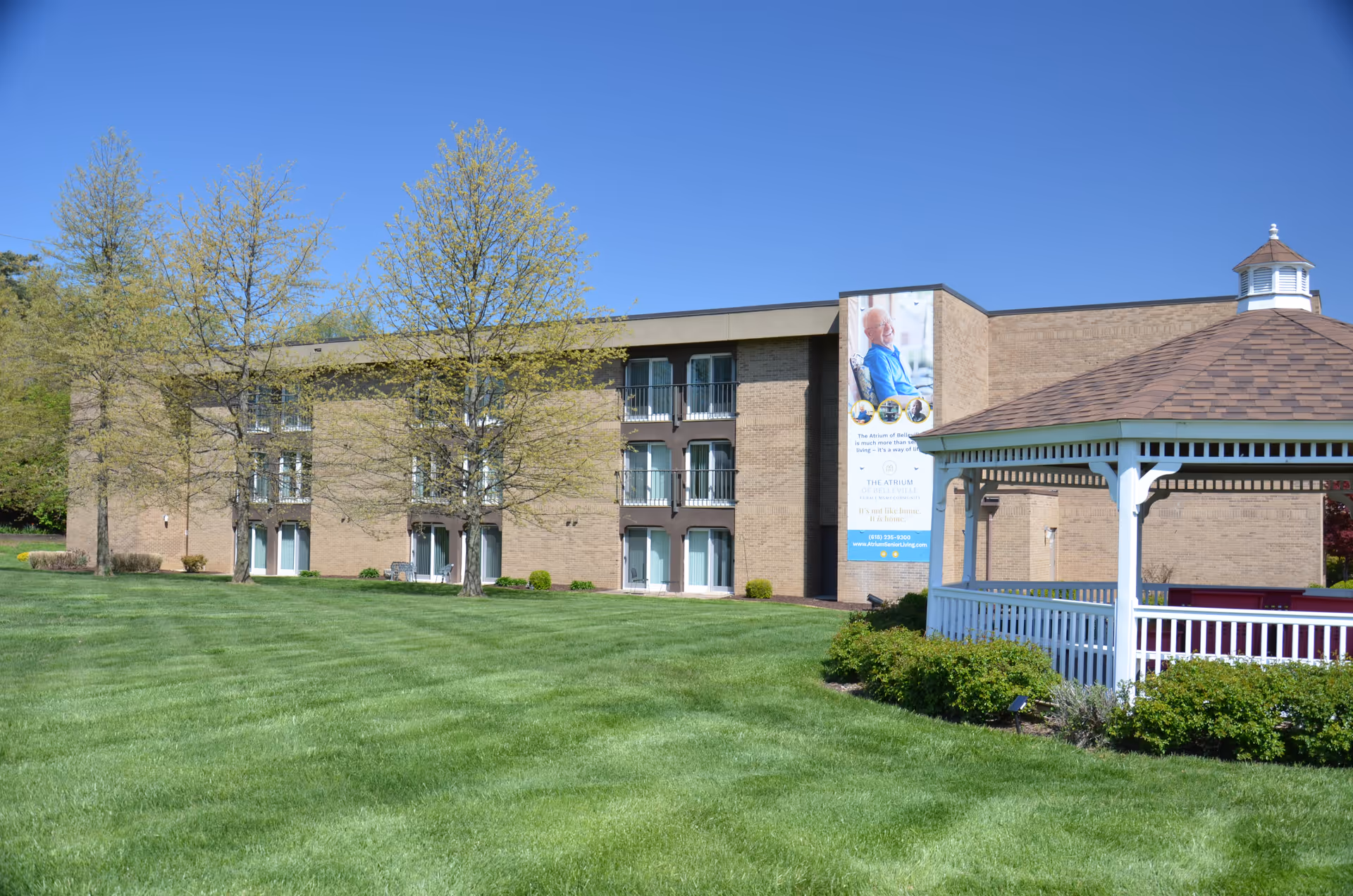 Exterior view of The Atrium of Belleville senior living facility on a clear sunny day. The building is three stories tall with large windows and balconies. In front of the building is a well-maintained green lawn with three trees that have light green leaves. To the right, there is a white gazebo with a brown shingled roof surrounded by bushes. A large vertical banner on the building shows an elderly man smiling and includes text about the facility.