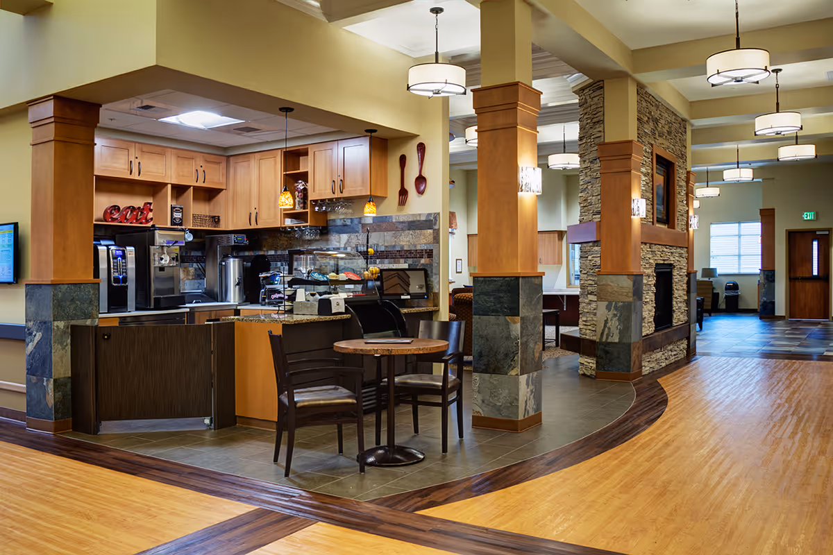 Interior view of a senior living facility showing a small kitchen area with wooden cabinets, coffee machines, and a display case with pastries. In front of the kitchen, there is a small round table with two chairs. The area has a mix of tile and wood flooring, stone pillars with wooden accents, and modern hanging light fixtures. A stone fireplace is visible in the background along with windows and doors.