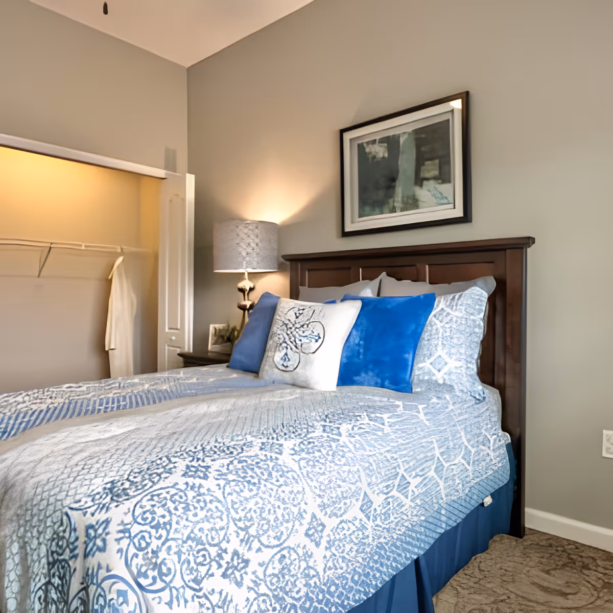 A neatly made bed with a blue and white patterned bedspread and several pillows in shades of blue and white. There is a wooden headboard, a bedside table with a lamp, and a framed abstract painting on the wall above the bed. An open closet with a hanging white robe is visible to the left.