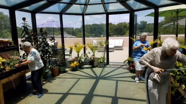 Three elderly women tending to plants inside a glass-enclosed sunroom with a view of an outdoor patio area with seating and umbrellas. The sunroom has a geometric glass ceiling casting shadows on the floor.