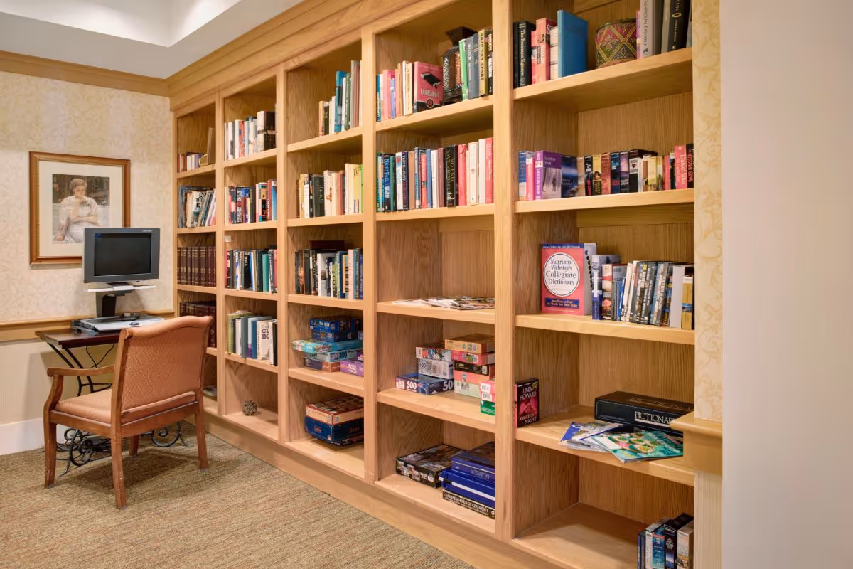 A cozy interior reading room with floor-to-ceiling wooden bookshelves filled with books and games, plus a small desk with a computer and chair.