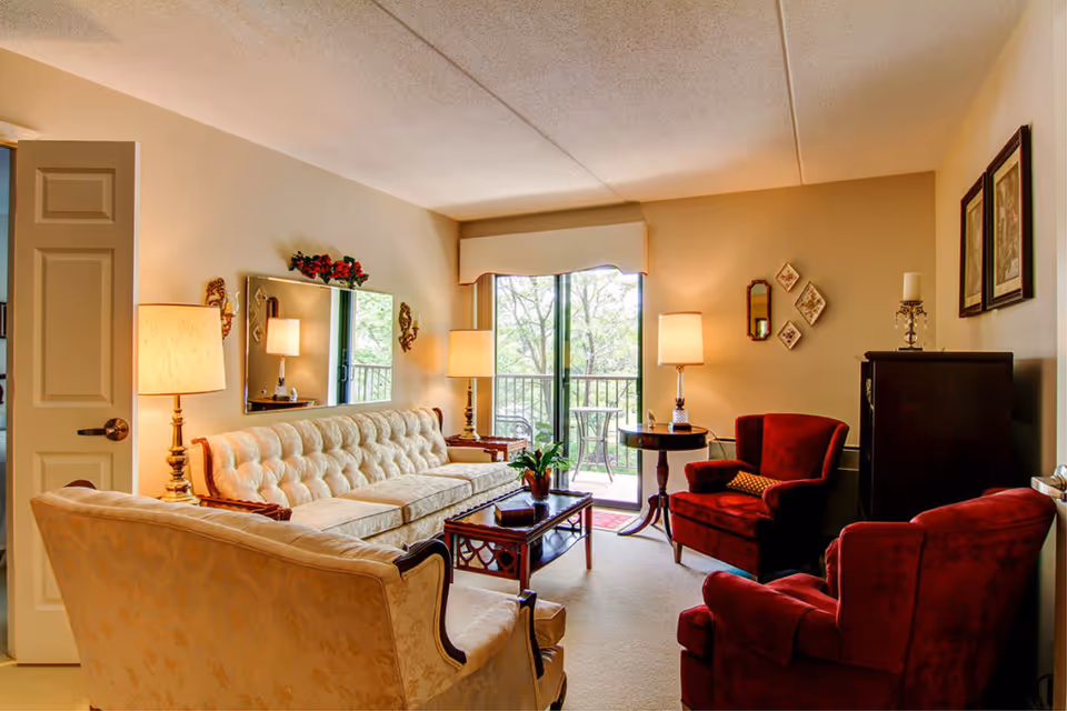 A cozy living room with a cream-colored tufted sofa, matching armchair, two red armchairs, a wooden coffee table with a plant, two side tables with lamps, a large mirror on the wall, and a sliding glass door leading to a balcony with outdoor seating and greenery visible outside.