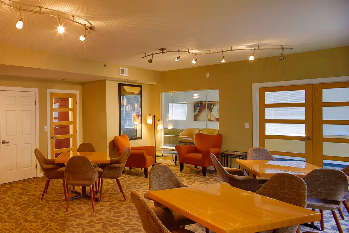 A well-lit common area with multiple wooden tables and cushioned chairs arranged around them. Two orange armchairs are positioned near a wall with a framed picture and a floor lamp. The room has beige walls, patterned carpet, and modern ceiling track lighting. There are glass-paneled wooden doors and a window reflecting another seating area.