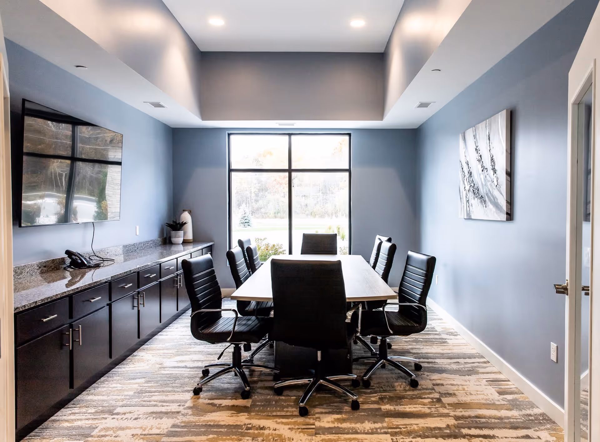 A modern conference room with a rectangular table surrounded by eight black office chairs on wheels. The room has blue-gray walls, a large window at the far end letting in natural light, a mounted flat-screen TV on the left wall, a long cabinet with a granite countertop beneath the TV, and a piece of abstract art on the right wall. The floor is covered with a patterned carpet.