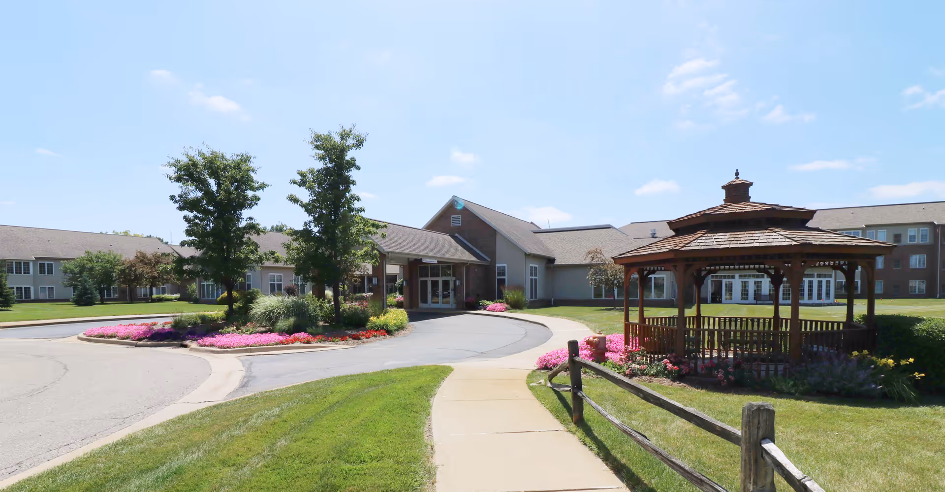 Front entrance of a senior living building with a circular driveway, manicured flower beds, and a wooden gazebo.