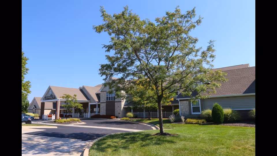 Exterior view of a senior living facility building with a driveway, green lawn, and a tree in the foreground under a clear blue sky.