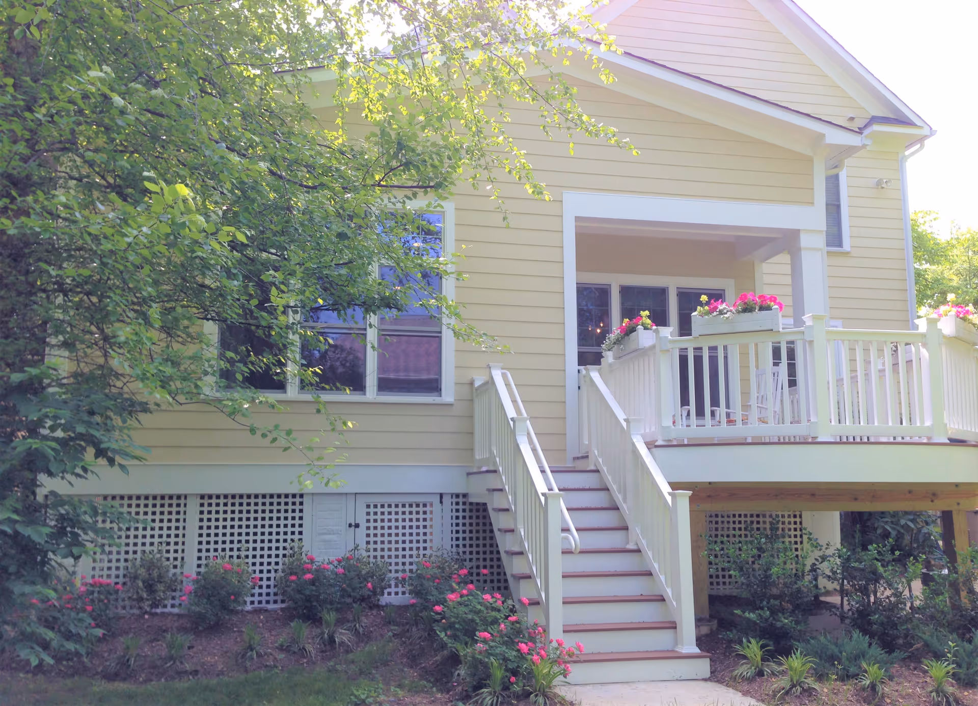 Front exterior of a yellow house with a white-railed porch, steps, and flowering plants.