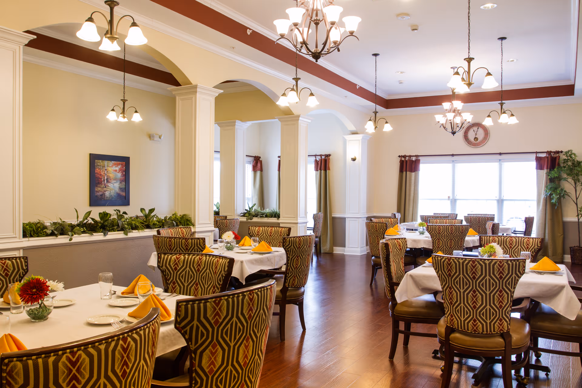 Bright dining room with multiple set tables, patterned chairs, chandeliers, and large windows.