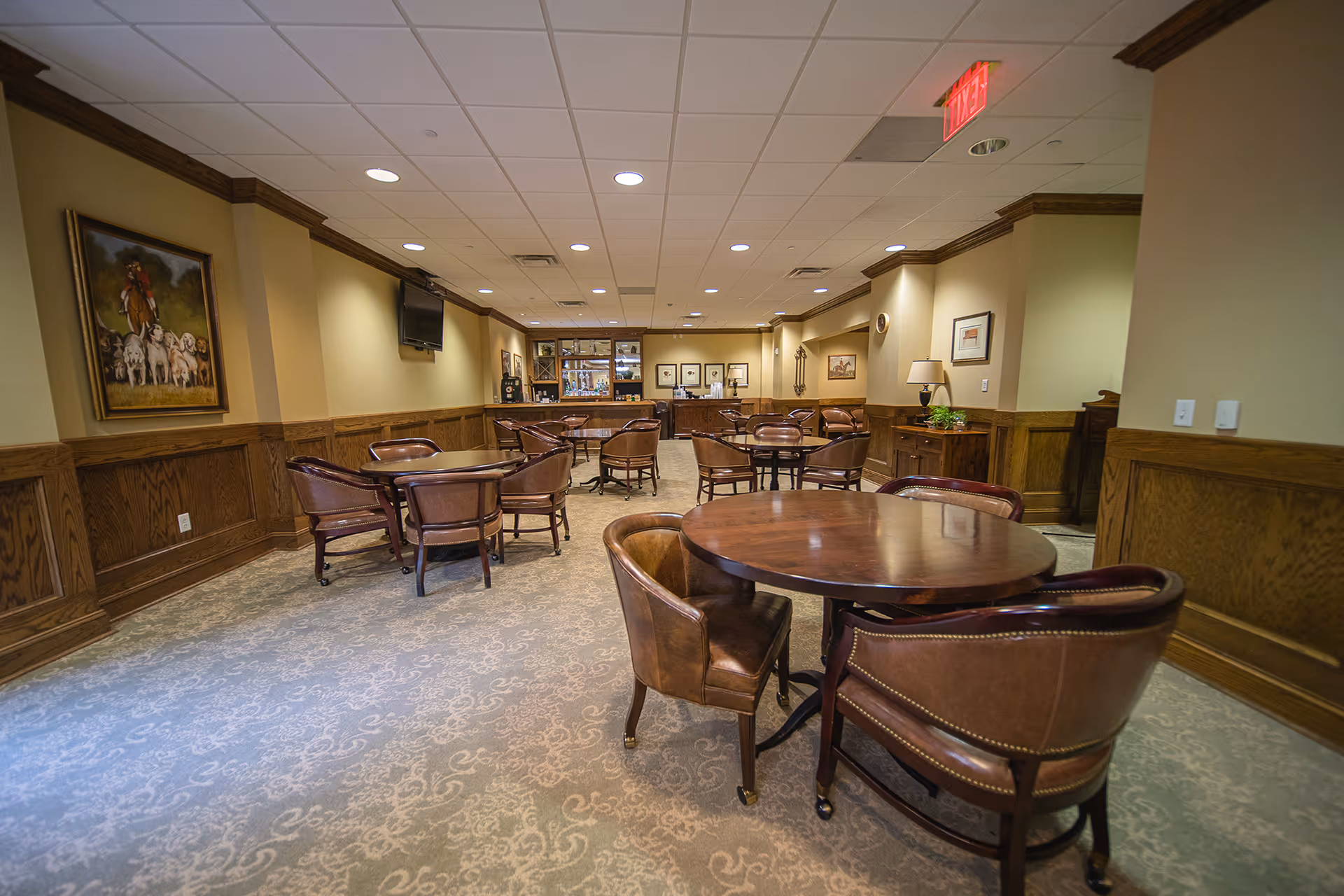 A spacious common area with multiple round wooden tables and brown leather chairs arranged around them. The room features wood paneling on the lower half of the walls, beige upper walls, and a patterned carpet. There are framed pictures and a painting on the walls, a flat-screen TV mounted on one wall, and a counter with shelves and decorative items at the far end of the room. The ceiling has recessed lighting and an exit sign is visible.