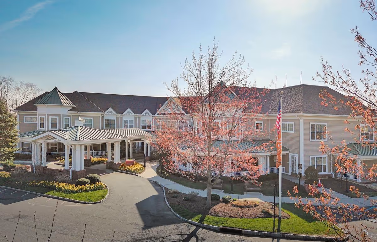 Exterior view of Sunrise of Woodcliff Lake, a large two-story senior living facility with a covered entrance, landscaped grounds with blooming trees and flower beds, and a flagpole with an American flag and an orange flag.