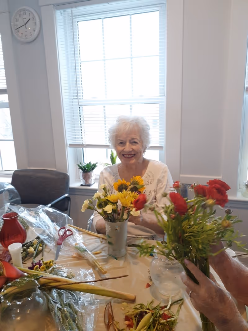 An elderly woman with white hair smiling while arranging flowers in a vase at a table covered with a plastic tablecloth. Various flowers, scissors, and vases are on the table. The room has large windows with white blinds and a clock on the wall showing 12:10.