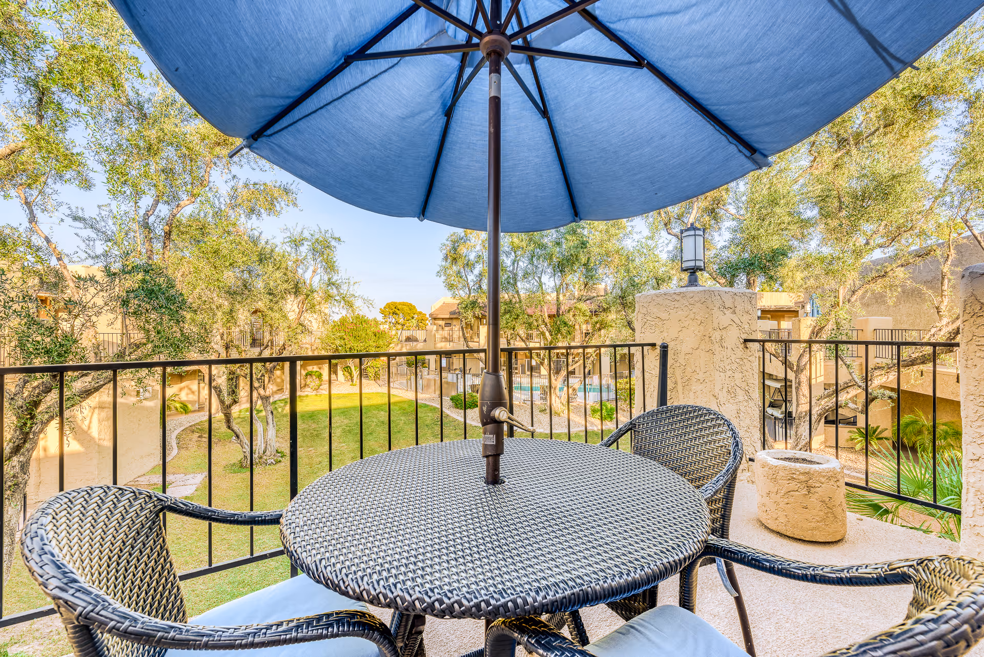 Outdoor patio area with a round wicker table and four matching chairs with cushions, shaded by a large blue umbrella. The patio overlooks a green lawn with trees and a swimming pool in the background, surrounded by beige stucco buildings and black metal railings.